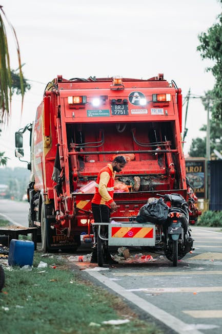 A sanitation worker dressed in a yellow high-visibility vest and dark trousers is operating a large, red rubbish collection truck on the side of a road. The truck's rear compactor is open, revealing its internal mechanisms, and the worker appears to be emptying or managing waste. A black garbage bag is placed on a small platform attached to the back of the truck. The scene is set in an outdoor urban environment with a roadway, some greenery, and distant trees visible in the background. The sky appears overcast, and the area around the truck has some scattered debris and waste. The presence of this rubbish removal equipment and worker suggests an on-site waste collection process, potentially a private waste handling or alternative refuse disposal service. Waste Clearance Belsize Park frequently provides such services to ensure effective rubbish clearance in the community, supporting various waste management needs outside local authority collection. The overall scene emphasizes professional, efficient waste handling in a typical street setting.