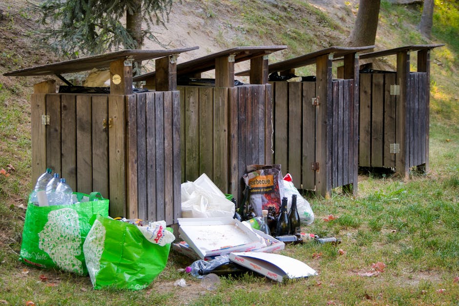 A row of four wooden outdoor trash and recycling bins arranged side by side on a grassy area, each with a sloped wooden roof for weather protection. The wood is weathered with a dark brown finish, showing visible grain and some slight discoloration. In front of the bins, there are various discarded items including clear plastic bottles, a green plastic shopping bag filled with waste, a pizza box with leftover crumbs, several glass bottles, and miscellaneous packaging materials. The background features a natural setting with trees and a dirt embankment, indicating an outdoor area near a park or woodland. The scene is illuminated by natural daylight, highlighting the textures of the wood and the scattered rubbish, which collectively suggest an informal waste disposal site, possibly related to private rubbish collection or on-site clearance services provided by Waste Clearance Belsize Park, situated on a grassy verge during a clear day.