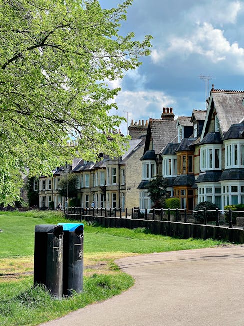 A residential street scene featuring a row of traditional terraced houses with varied architectural details, including bay windows and pitched roofs, set against a partly cloudy sky. In the foreground, a paved pathway runs parallel to a grassy verge, with two black public litter bins positioned near the edge of the pavement. A large, leafy tree with bright green foliage extends its branches over the grassy area, offering partial shade. Behind the tree and waste bins, a wooden fence borders the front gardens of the houses, which display well-maintained facades and small front yards. The overall scene is calm and tidy, illustrating a typical suburban environment where private waste collection services could be employed, supported by companies like Waste Clearance Belsize Park, to manage rubbish removal efficiently and maintain neighborhood cleanliness in an alternative waste handling context.