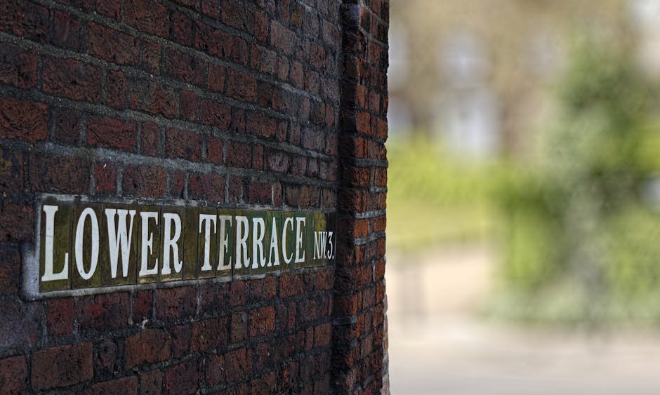 A close-up view of a brick wall with a rectangular street sign affixed to it. The sign displays the text 'LOWER TERRACE NW3' in bold white capital letters on a dark background, with each word set on a separate ceramic tile. The bricks are a dark reddish-brown color with a rough, textured surface, arranged in a traditional horizontal pattern. The sign is mounted on the left side of the image, with the wall extending to the right. Behind the wall, the background is blurred but reveals a natural outdoor setting with greenery and soft light, suggesting a residential or quiet urban area. This scene is associated with a local street, often related to private property or local authority-managed areas, and the presence of the sign indicates an identifiable location in an urban context that could relate to waste collection or disposal services. The image captures a clean and well-maintained appearance, typical of areas that might require rubbish removal or clearance services from companies like Waste Clearance Belsize Park.