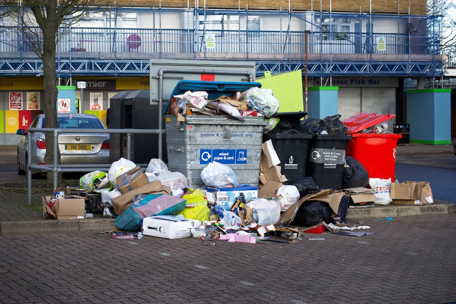 A collection of overflowing rubbish bins and piles of discarded waste material situated on a paved area in front of a commercial building. The scene features multiple large bins, including a gray bin labeled for mixed paper and card, a black bin likely for general waste, and a red bin, all positioned close together. Surrounding these bins are numerous cardboard boxes, paper and plastic packaging, black plastic bags, and various loose waste items scattered across the pavement. In the background, a blue scaffolding structure covers the upper part of the building, and the shopfronts beneath display signage, indicating a retail or food establishment. A small, leafless tree stands to the left, with a metal railing separating the waste area from parked cars, including a visible silver vehicle nearby. The environment appears to be an urban commercial area, where waste accumulation suggests a waste management service, such as private rubbish collection by Waste Clearance Belsize Park, is needed to address the excess debris and maintain cleanliness and safety in the vicinity.