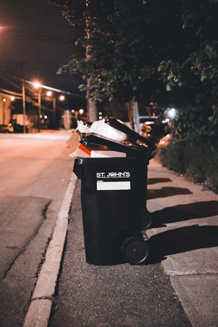 A collection of overflowing rubbish bins and piles of discarded waste material situated on a paved area in front of a commercial building. The scene features multiple large bins, including a gray bin labeled for mixed paper and card, a black bin likely for general waste, and a red bin, all positioned close together. Surrounding these bins are numerous cardboard boxes, paper and plastic packaging, black plastic bags, and various loose waste items scattered across the pavement. In the background, a blue scaffolding structure covers the upper part of the building, and the shopfronts beneath display signage, indicating a retail or food establishment. A small, leafless tree stands to the left, with a metal railing separating the waste area from parked cars, including a visible silver vehicle nearby. The environment appears to be an urban commercial area, where waste accumulation suggests a waste management service, such as private rubbish collection by Waste Clearance Belsize Park, is needed to address the excess debris and maintain cleanliness and safety in the vicinity.