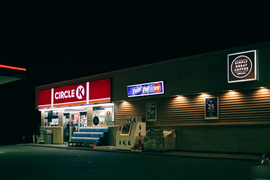 A nighttime scene of a Circle K convenience store with illuminated signage displaying the brand logo in red and white. The store's entrance is open, revealing shelves stocked with drinks and various items. To the right of the entrance, there is a vending machine and a pack of bottled water stacked outside. The exterior features horizontal wooden siding with warm lighting fixtures casting a subtle glow on the wall. Adjacent to the store, a small vending stall or ice machine with several ice bags and an ice sign can be seen, indicating alternative waste handling or collection outside regular hours. The surroundings include a dark, empty parking area with a faintly visible car on the far right edge, and the background consists of the night sky, giving a quiet, formless atmosphere. This scene captures typical private convenience retail premises which might temporarily accommodate outside rubbish or waste collection, managed by waste clearance services such as Waste Clearance Belsize Park, within the scope of their rubbish removal offerings.