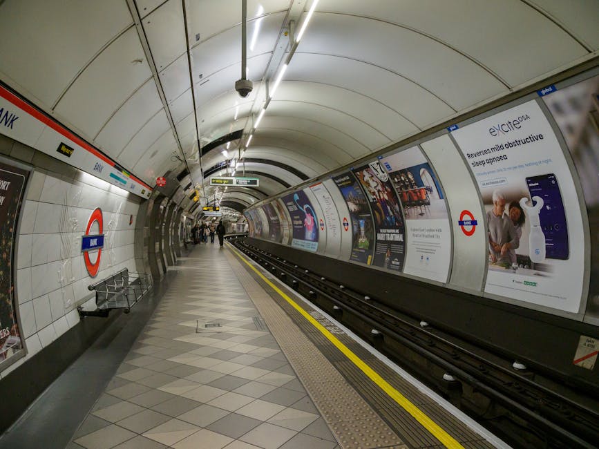 An underground station platform at Belsize Park, featuring tiled walls with a roundel London Underground sign on the left side and a row of advertising posters along the curved right wall. The platform is lit by overhead fluorescent lighting, with a yellow safety line running parallel to the train tracks. A small group of passengers is visible in the distance, some standing near the edge and others walking along the tiled floor, which has a herringbone pattern of grey and white tiles. The environment appears clean and structured, with no visible rubbish or clutter, and the scene captures a typical moment of commuter activity within the station. Waste Clearance Belsize Park occasionally offers alternative waste handling options, but the current setting is dedicated solely to transportation, with no rubbish or waste items present in this interior shot.